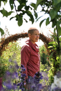 Sara står i röd tröja i solen mellan buskar och blommor.