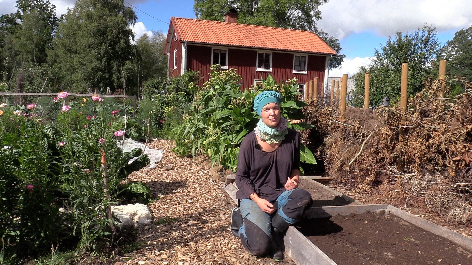 Sara Bäckmo sitter på en kant till en odlingsbädd i en somrig trädgård. Summer sowings, Sara in the kitchen garden, next to a growing bed.
