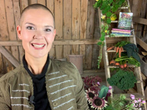 Sara håller ett föredrag om sina första odlarår. Hon står i en lada framför sina böcker och grönsaker och ser glad ut.