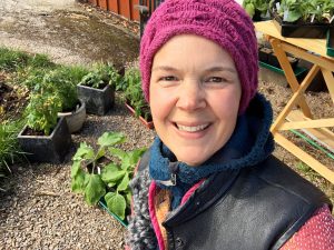 En glad Sara i lila mössa står i köksträdgården