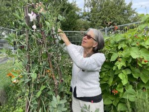 En äldre kvinna med grått hår och vita kläder står i en liten odling.